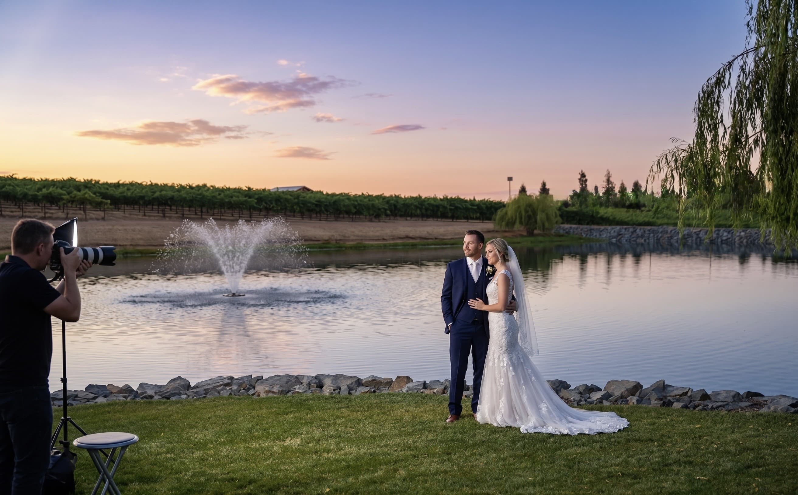 A couple in wedding attire being photographed at the edge of the lake at sunset, the fountain playing and the vineyard beyond