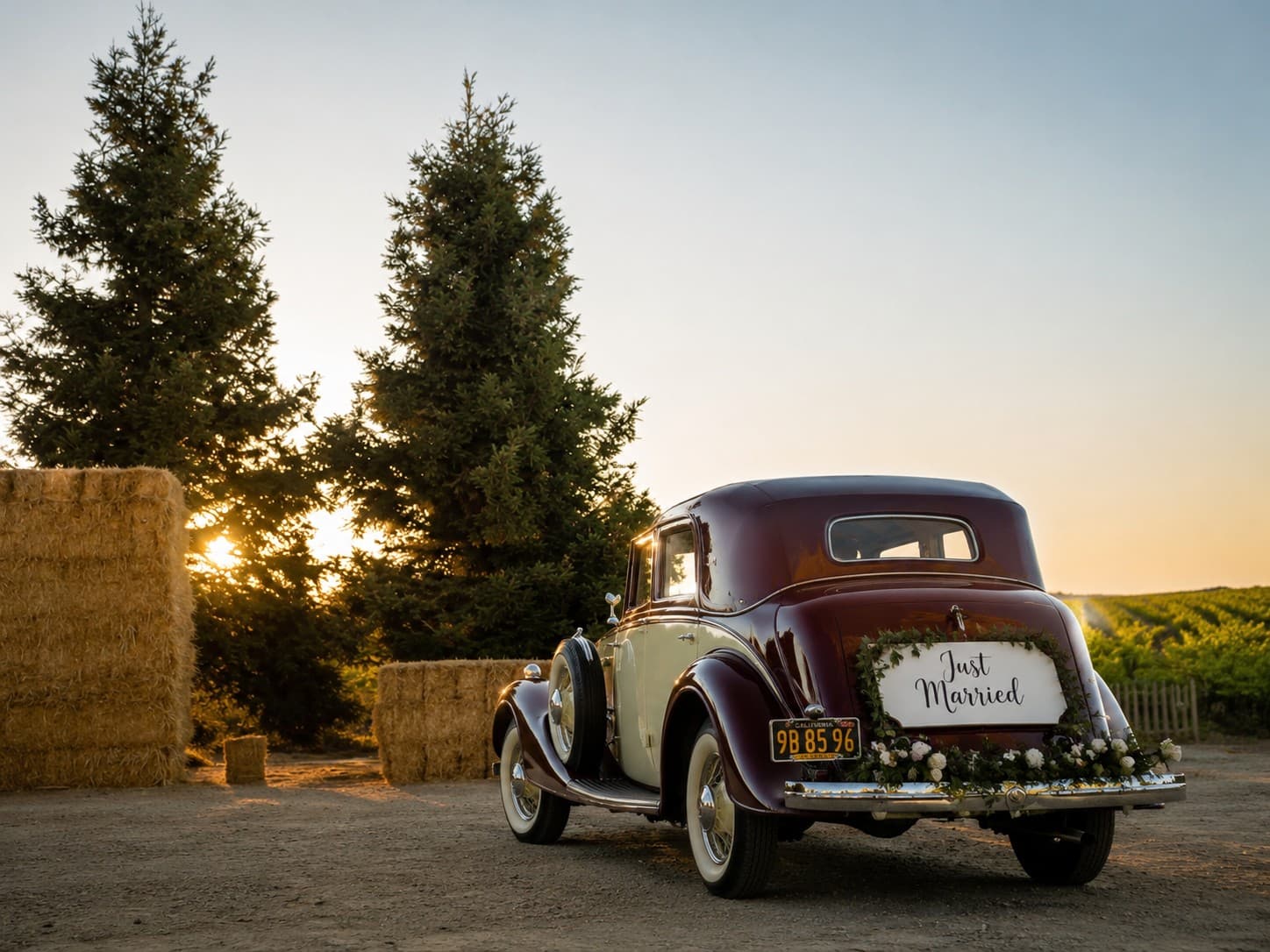 A classic getaway car marked Just Married at sunset, hay bales and the vineyard behind it